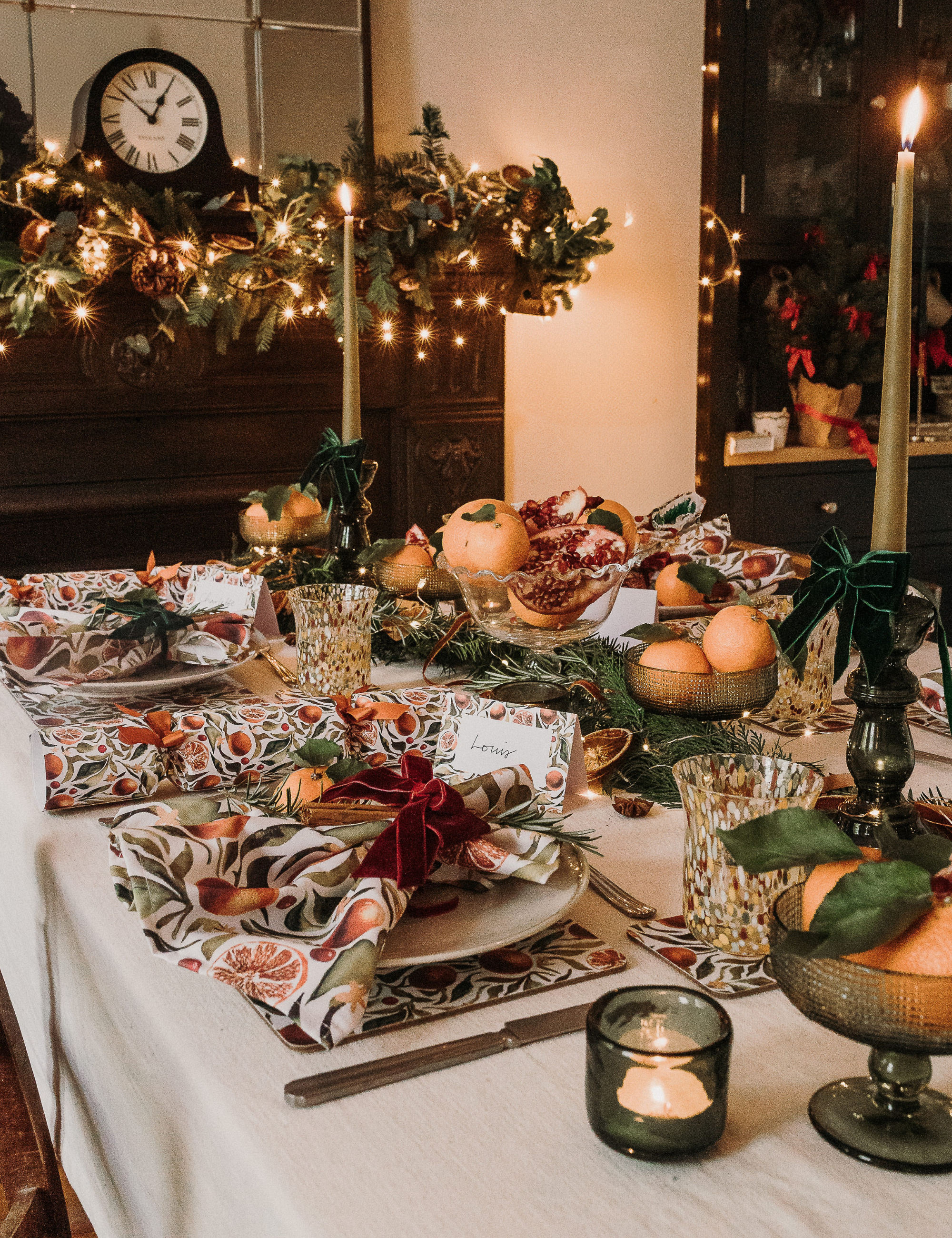 Charming Christmas tablescape featuring fruit-patterned table linens, velvet shoes, and tall moss green taper candles.