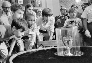 children in a museum look at a small, dark rock encased in a protective glass container