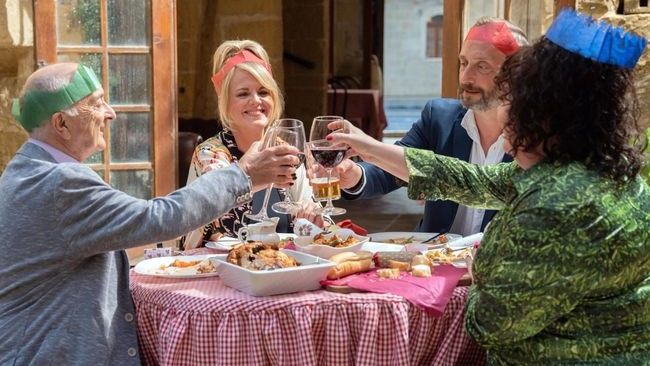 Patrick (Tony Robinson), Jean (Sally Lindsay), Dom (Steve Edge) and Gloria (Sue Vincent) sit around a table outdoors, toasting with glasses of wine. Each of them is wearing a paper hat from a Christmas cracker, and there is a Christmas dinner on the table in front of them.