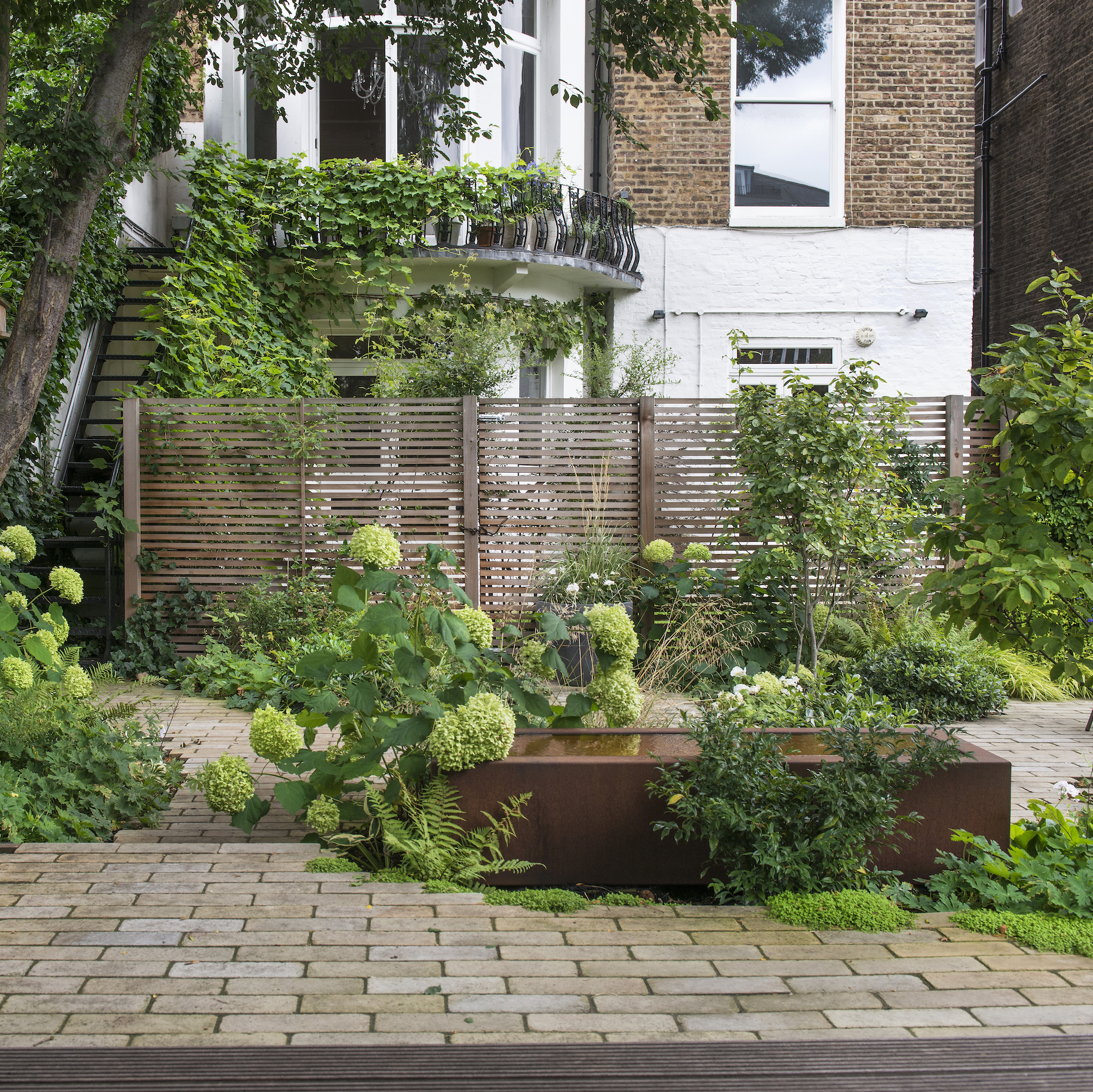 Terraced garden with green plants, metal water feature and wooden fence