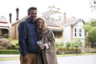 A shot from Colin From Accounts showing Gordon (Patrick Brammall) and Ashley (Harriet Dyer) standing outdoors. They are both smiling; Ashley has her right arm around Gordon's waist and is holding a plastic bag of dog treats in her other hand.