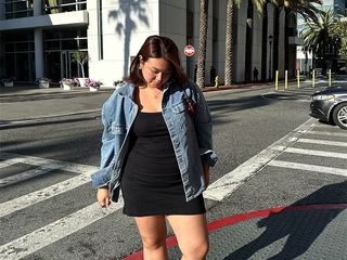 marina wears black mini dress, denim jacket and butter yellow sandals while standing on a street corner