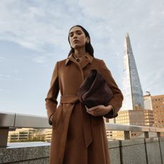 Model wearing a light brown wool coat holding a brown suede bag sold at Hobbs in front of the Shard