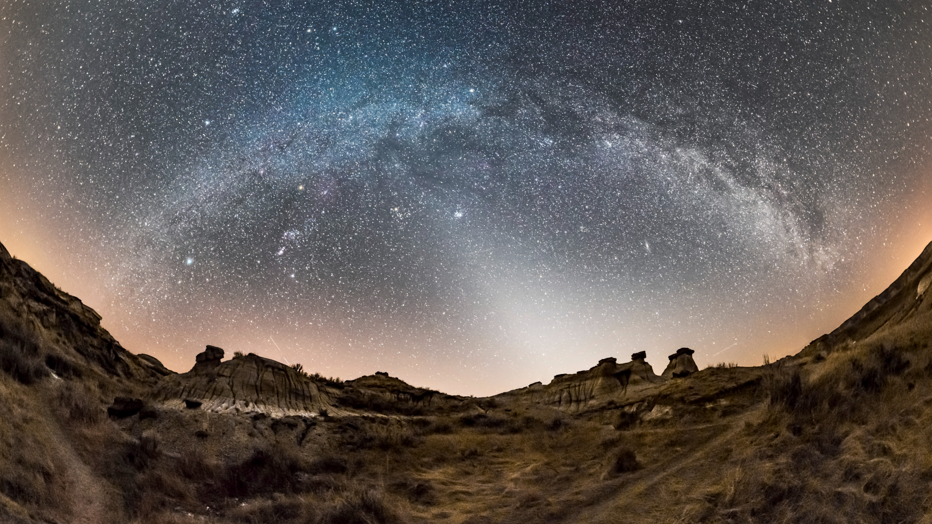 The bright glowing band of the Milky Way arcs over an arid, rocky land in a sky filled with stars.