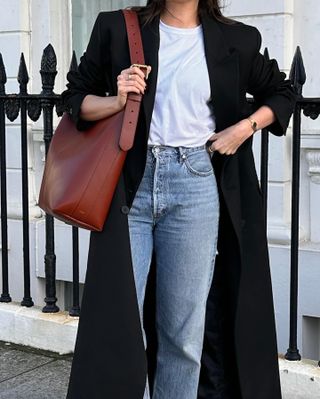 British fashion influencer Anna Newton poses in a black maxi coat, white t-shirt, leather bag, and straight-leg jeans.