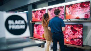 Two people in a retail store standing in front of several TVs available for sale. In the foreground is a sign that reads: "Black Friday."