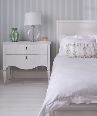 A white and gray bedroom with striped wallpaper, a nightstand with a glass lamp on it, and a white bed with a beige headboard and ruffled white bedding, with a gray wooden floor underneath