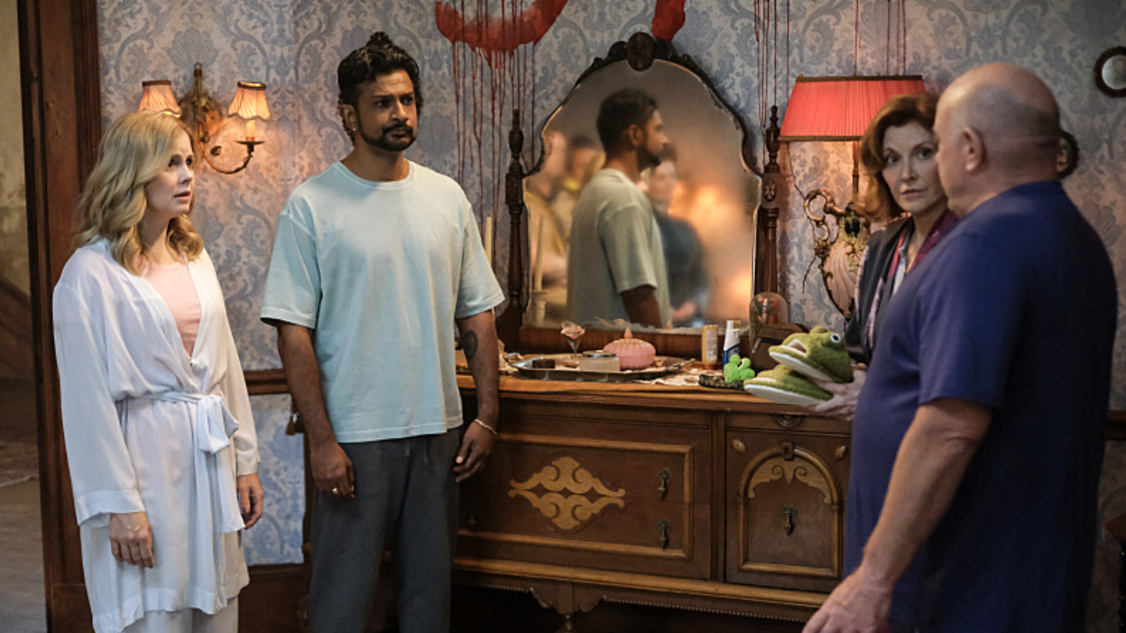 Dressed in pyjamas, Frank (Dean Norris) talks to Sam (Rose McIver), Jay (Utkarsh Ambudkar) and his girlfriend, as blood drips down the walls and ghosts are visible in the mirror in Ghosts US season 4