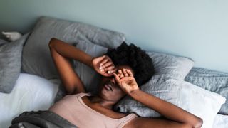 A woman lies in bed in the morning rubbing her eyes as she lays on gray and white bedding.