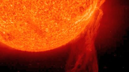 A close up of the sun shows a glowing orange and red ball of gas with long streaks of gas filaments leaking from its surface into the darkness of space.