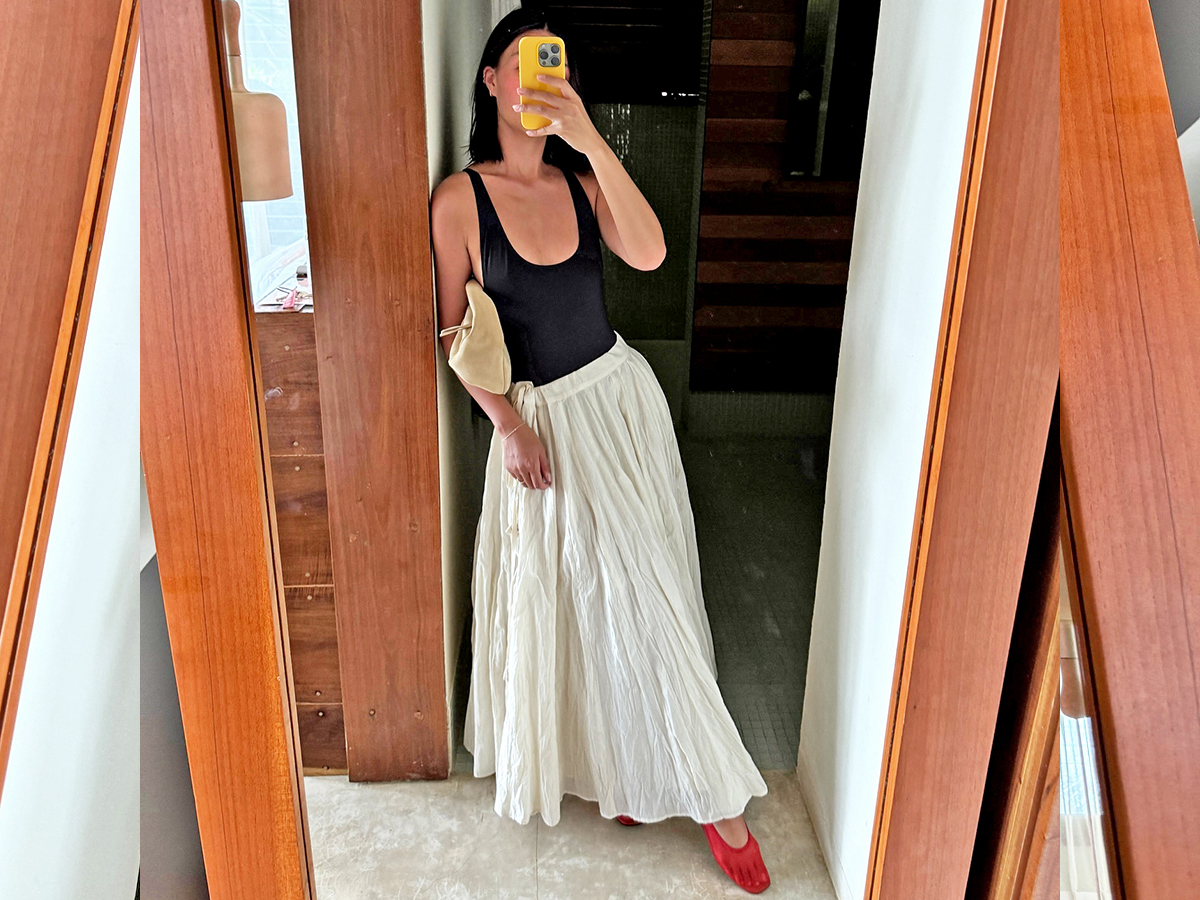 fashion influencer Mimi Nguyen poses for a mirror selfie while on vacation in Tulum Mexico with a yellow iphone case wearing a low-cut black tank top, tan suede bag, full white maxi skirt, and red mesh flats