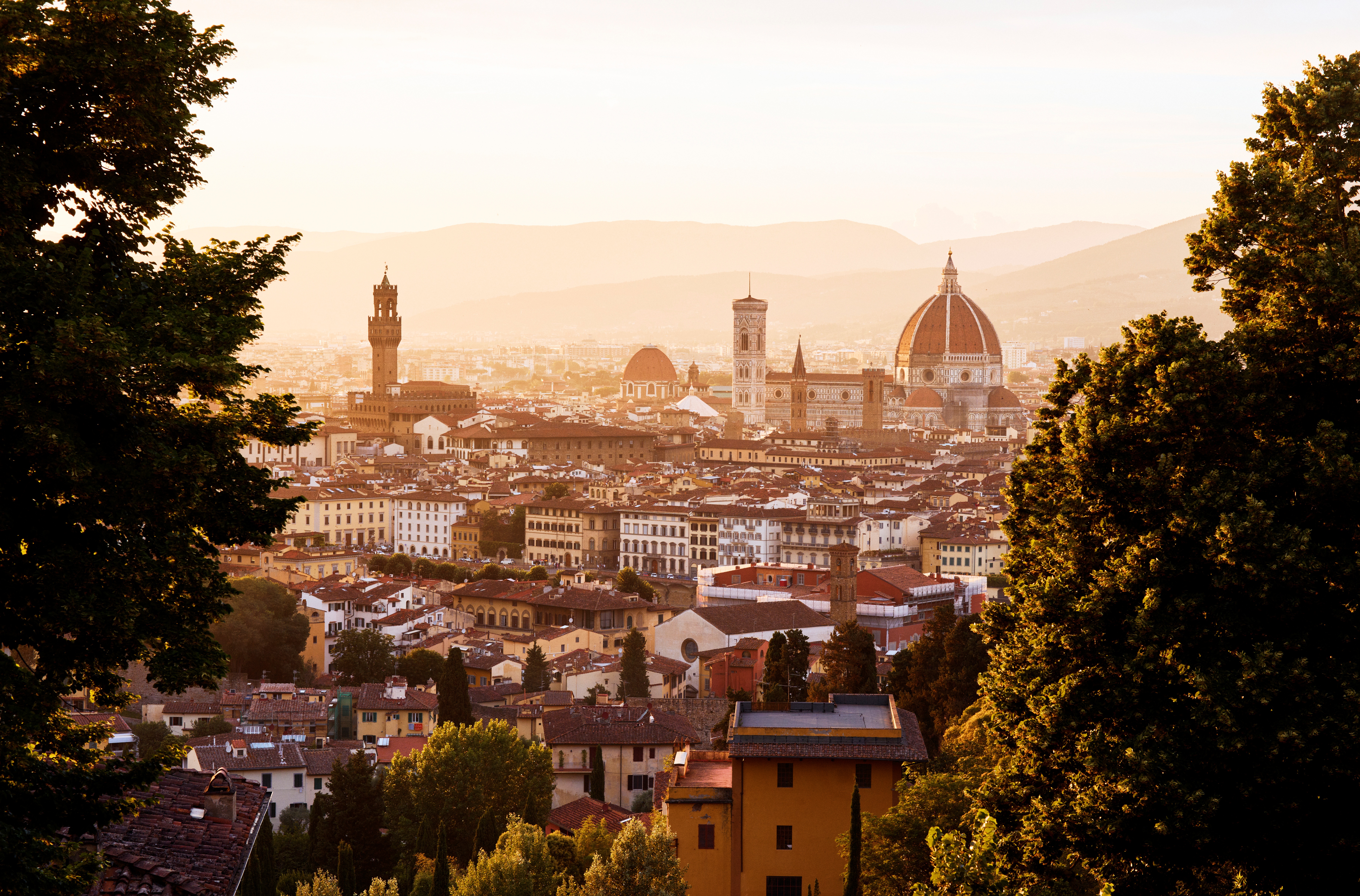 A city captured at sunset on a dusk-filled day shows historical buildings, mountains, and plants covered in fading sunshine.