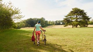 Woman pushing bike through grassy field, holding helmet, in the sunshine
