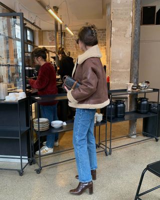 French influencer wearing brown motto jacke,t blue jeans, and brown ankle boots while holding a black Goyard bag and standing in coffee shop.