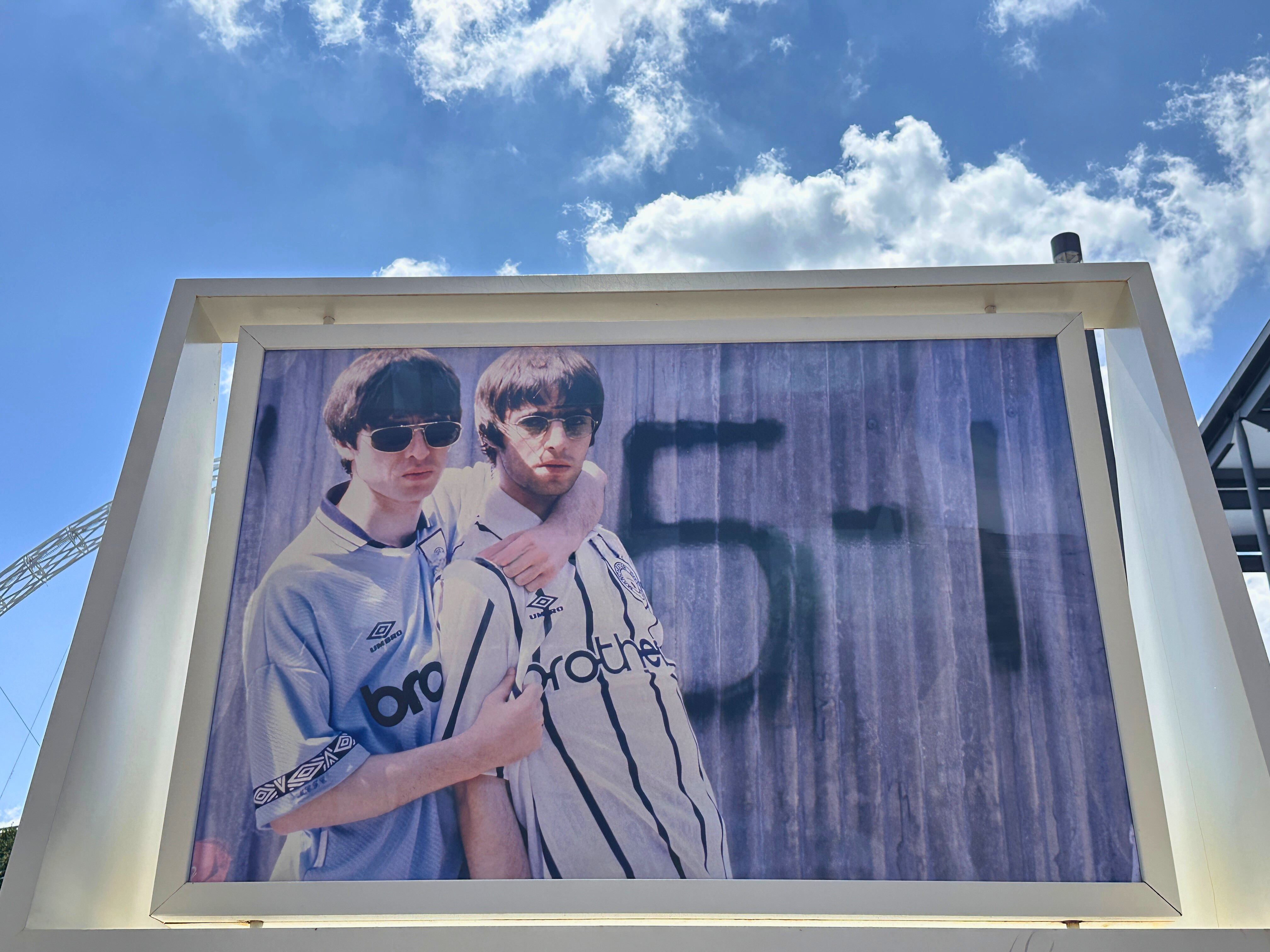 A large-scale photograph of Oasis members Liam and Noel Gallagher embracing each other in front of a corrugated metal wall with the numbers &quot;5-1&quot; painted on it.