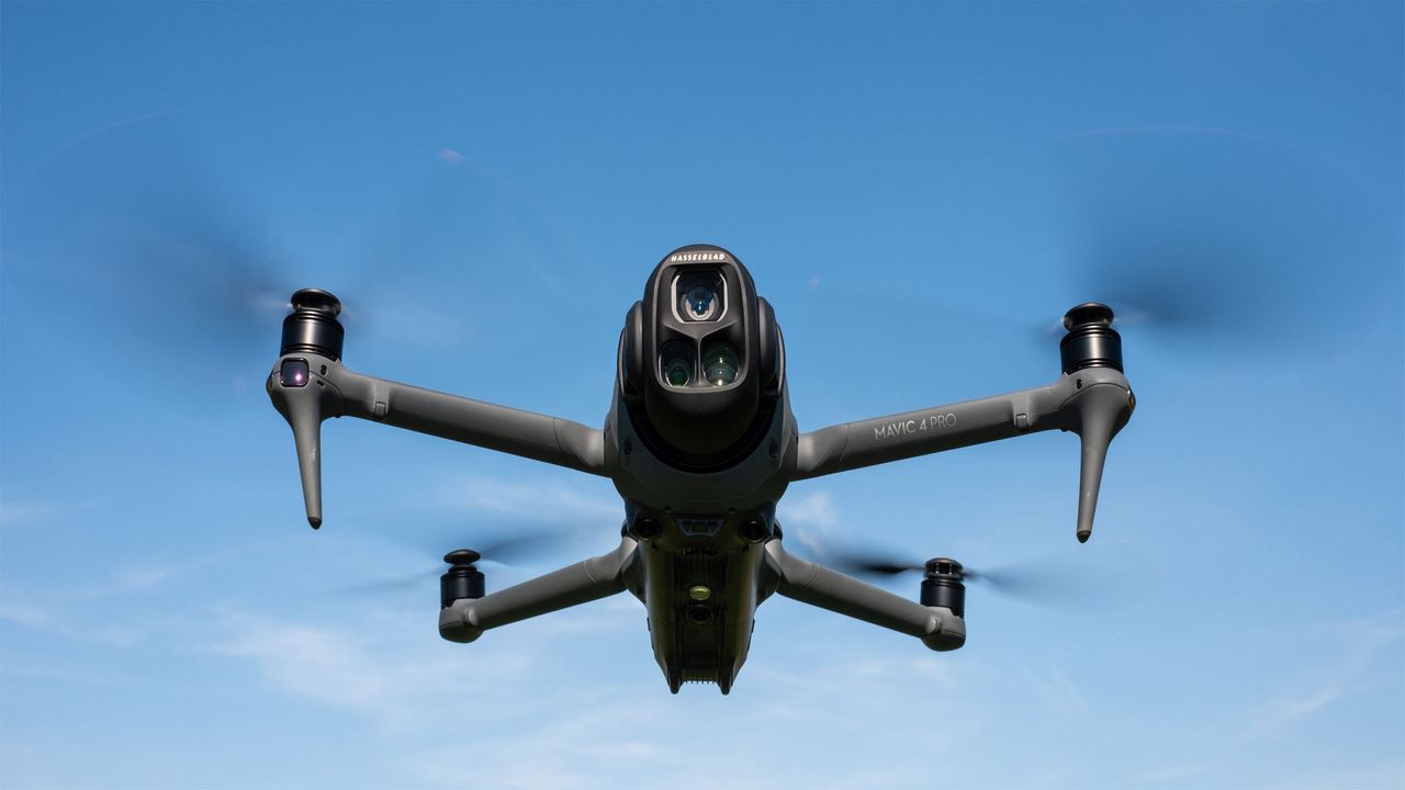 A dark-colored drone is mid-flight with its propellers in a blur on a clear blue sky background.