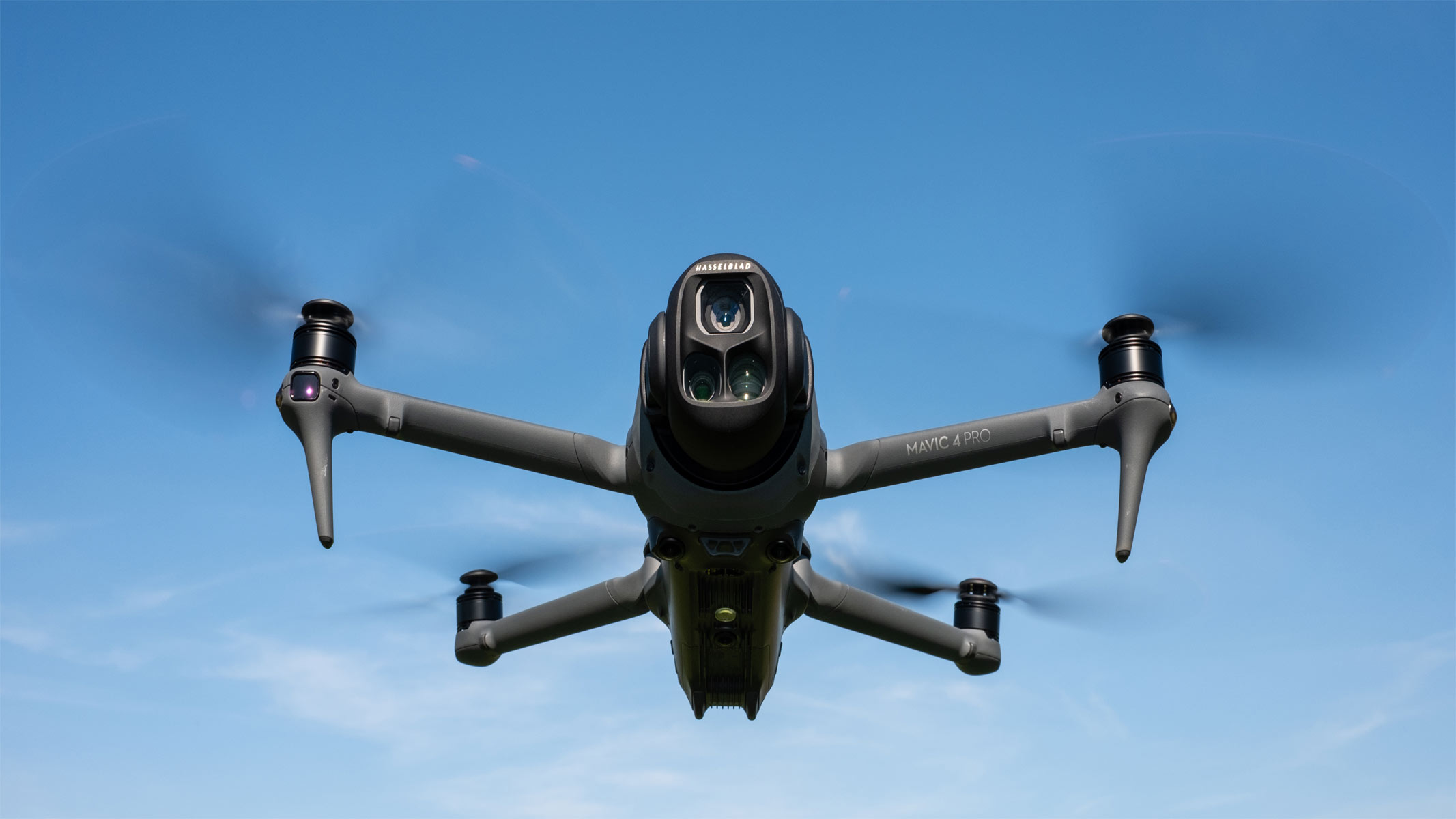 A dark-colored drone is mid-flight with its propellers in a blur on a clear blue sky background.