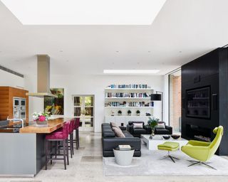 Kitchen with grey island unit and cupboards and plum coloured bar stools, with a relaxed seating area with black leather sofa and chairs, a modern green chair and matching footstool and a black fire surround with glass walls either side.