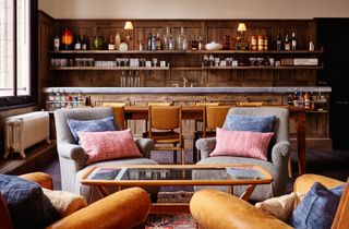 The Hoxton cocktail lounge with wooden paneled bar and grey marble top, mustard and grey armchairs with blue and pink cushions