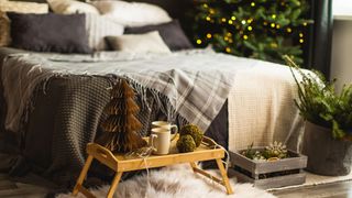 Image shows a cozy guest room dressed for Christmas, with plaid blankets on a bed and a wooden tray filled with earthen cups and green and burgundy paper decorations