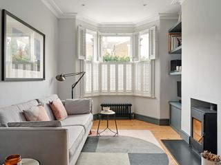 White living room with woodburning stove, light grey sofa, grey geometric rug and white shutters in a bay window