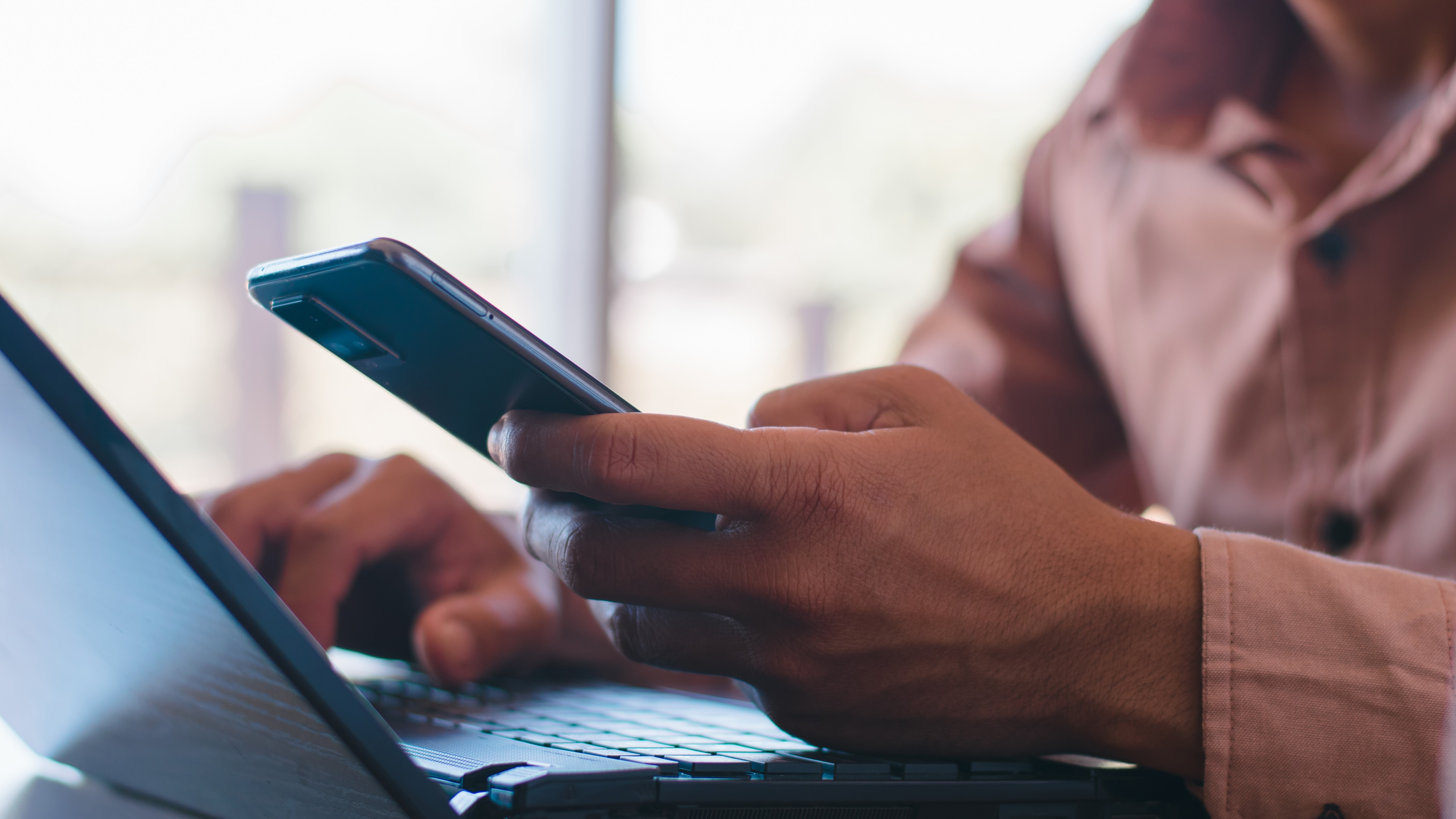 A man holding a smartphone while using a laptop, seemingly on a surface