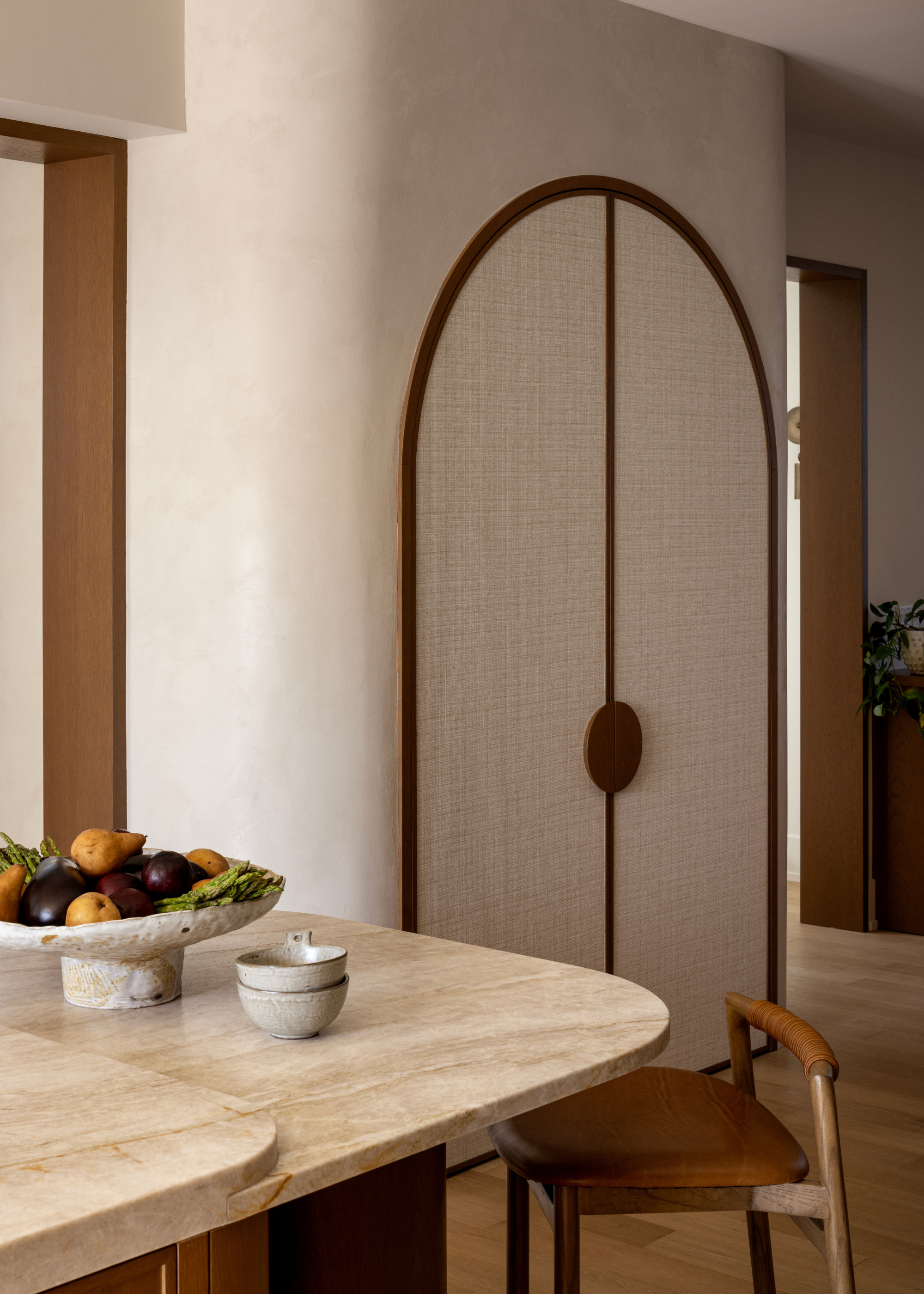 An open kitchen concept with a stone island holding a bowl of fruit by a storage closet with an arched top and a wood handle