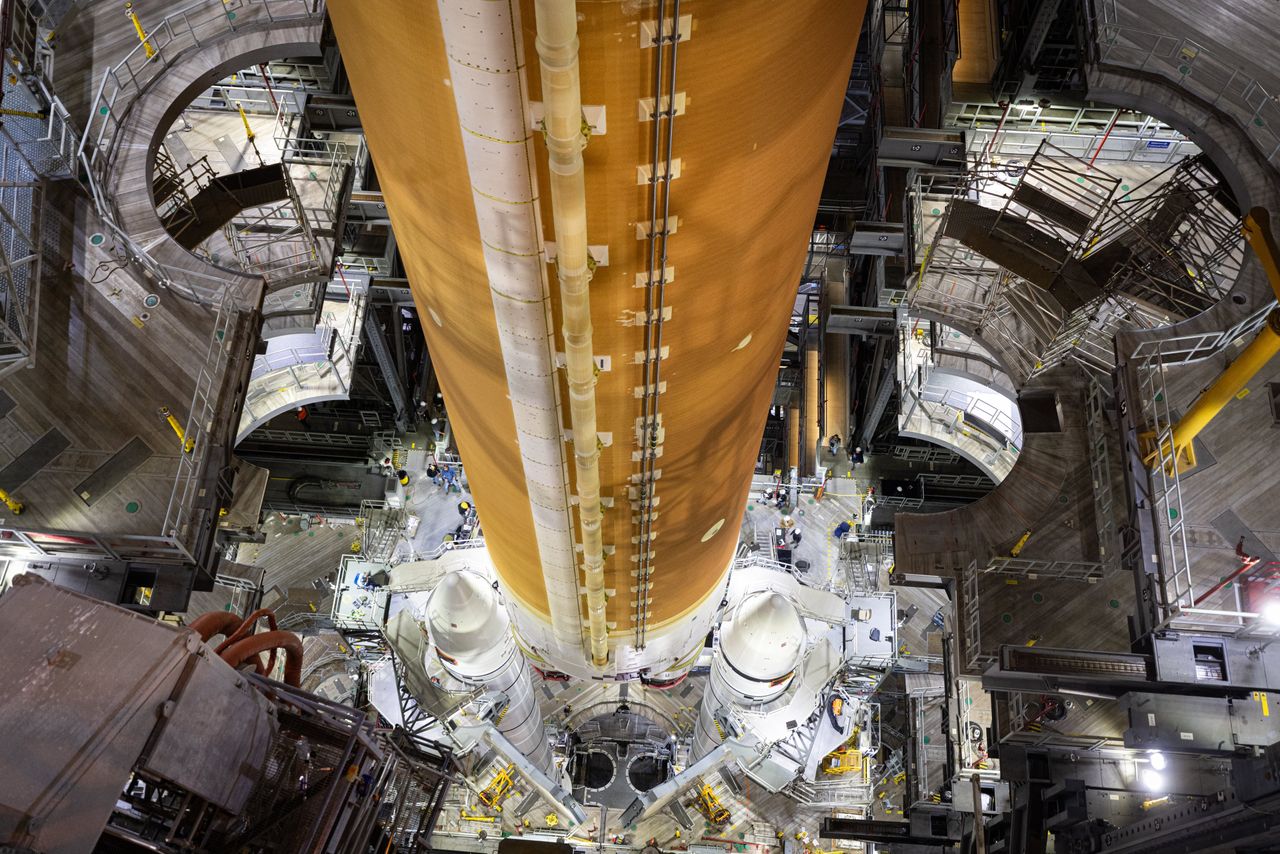 a large orange rocket booster is hoisted in a tall factory above two pointy white boosters secured by scafolding.