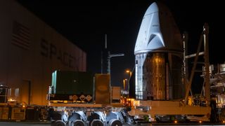 a white space captule with metalic bottom trunk rolls into a hangar on a truck at night time.