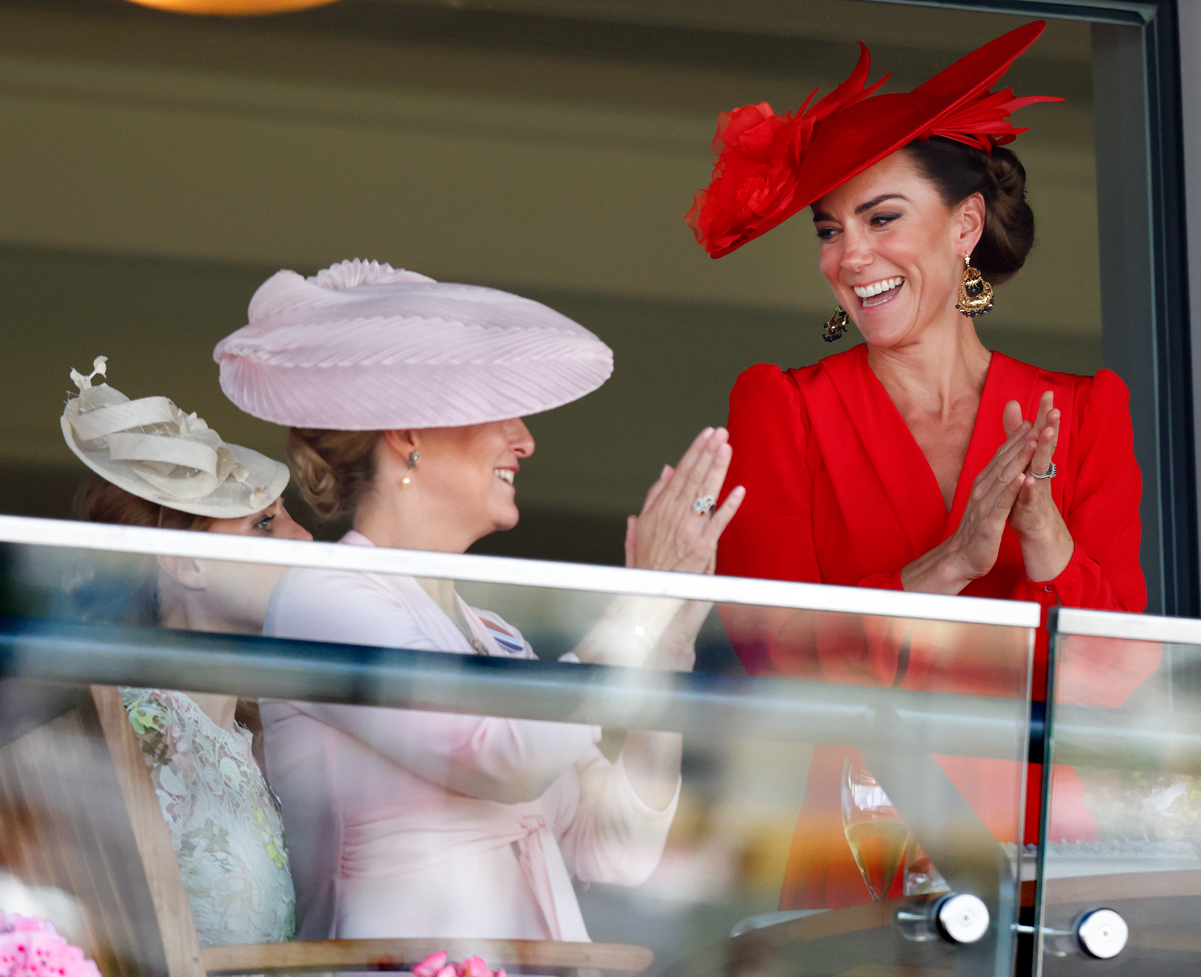 Duchess Sophie wearing a pink hat and clapping, smiling at Princess Kate, in a red dress and hat, at Royal Ascot