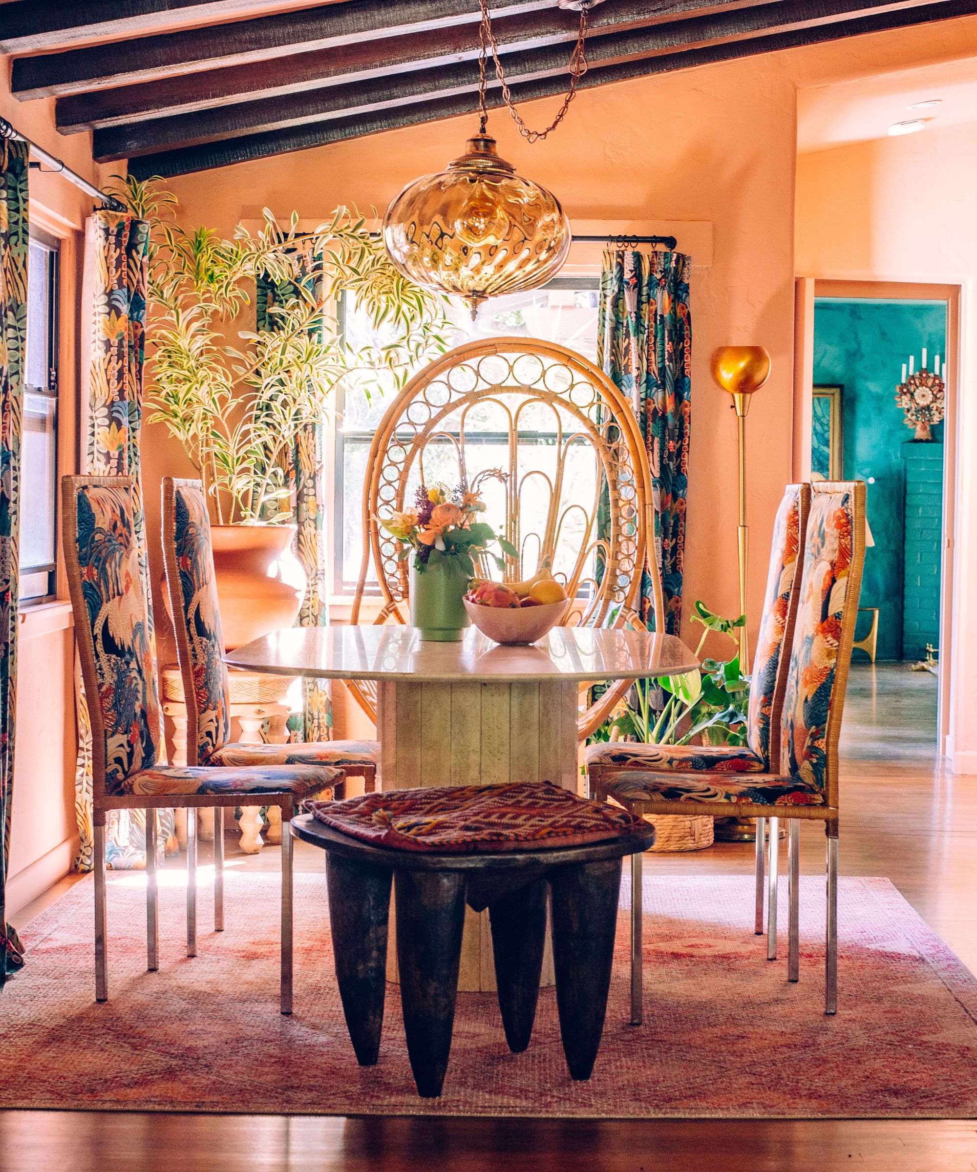 Dining room with peach walls, rattan armchair, round marble table, floral chairs, wooden stool and beamed ceiling with large potted plant and Moroccan light