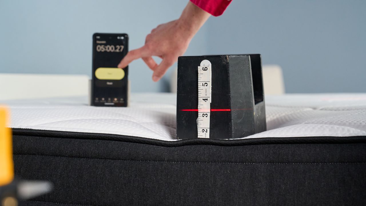 A close up of a 25kg weight perched on the edge of the Plank Firm mattress. A laser measure pointed at the weight shows it has sunk just over 3 inches. In the background, a hand reaches to stop the timer on a smartphone after 5 minutes