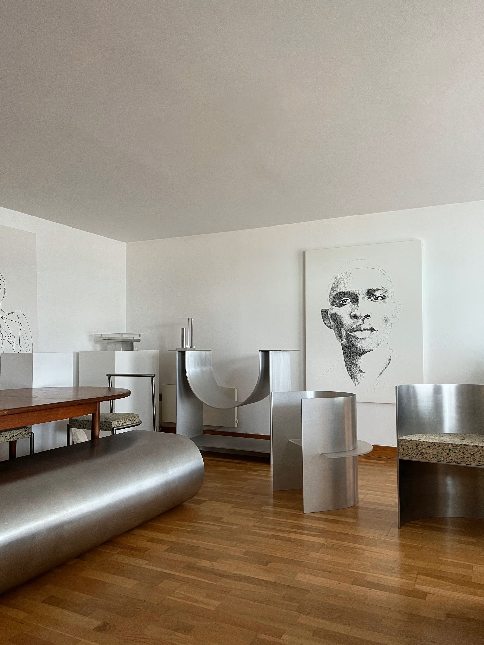 Sculptural, matte chrome furnishings, including chairs and banquettes, are styled in a wooden-floor living room with wood and chrome furniture, and a black portrait against a white canvas.