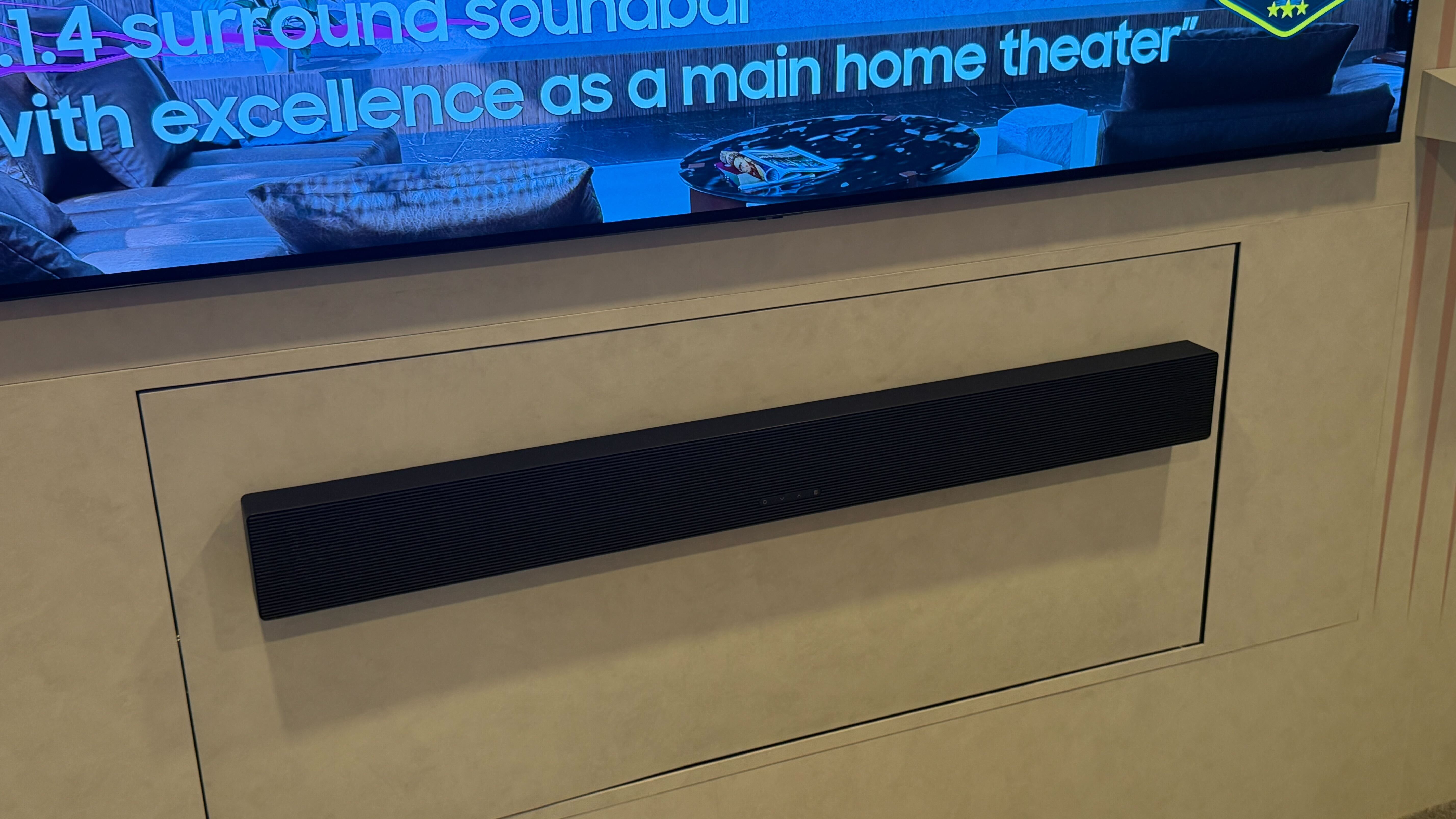 Samsung's HW-QS90H sitting below a wall-mounted TV on a white backdrop