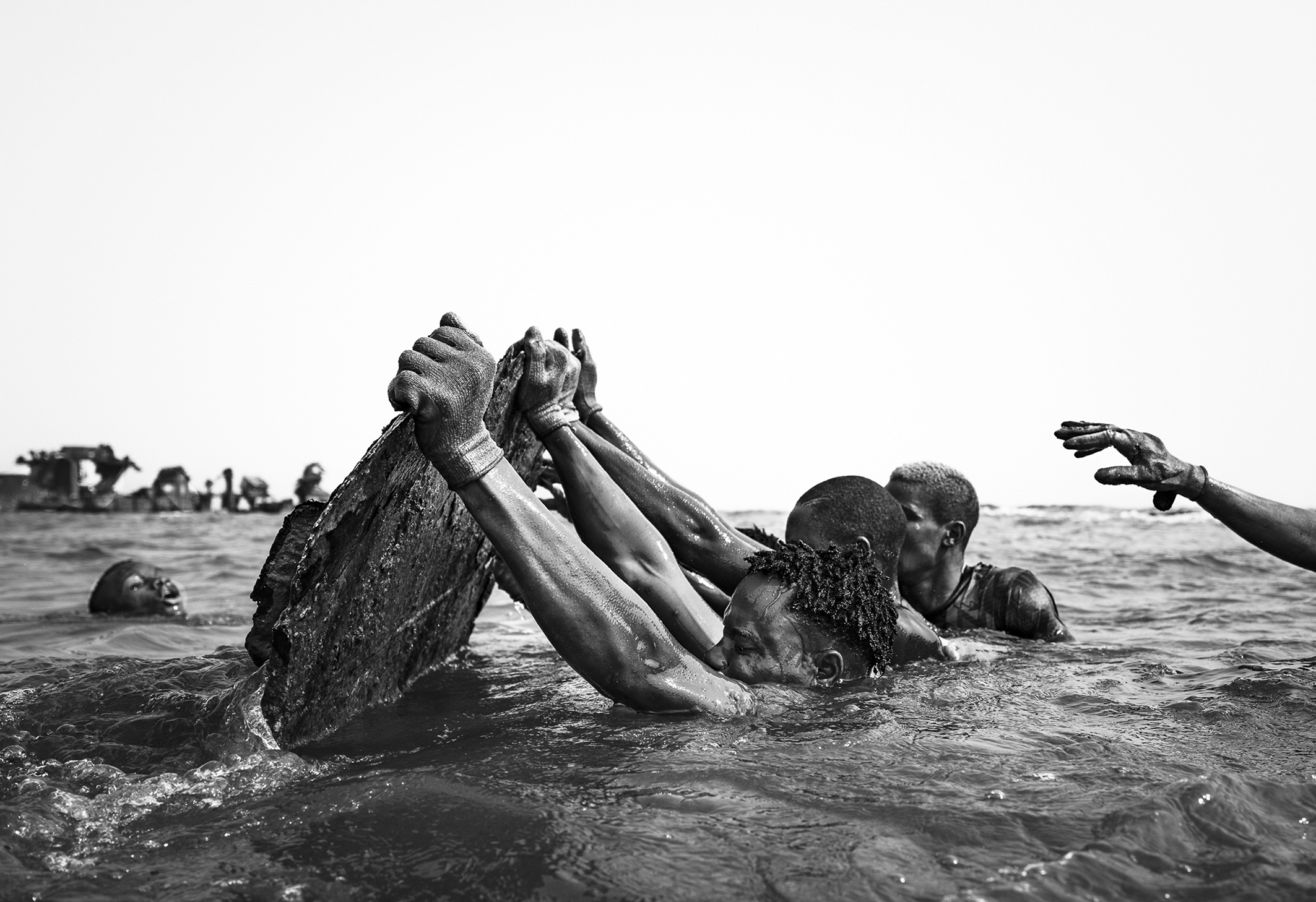 A group of people struggles to lift a heavy object from the water, showing determination and teamwork in a monochrome setting