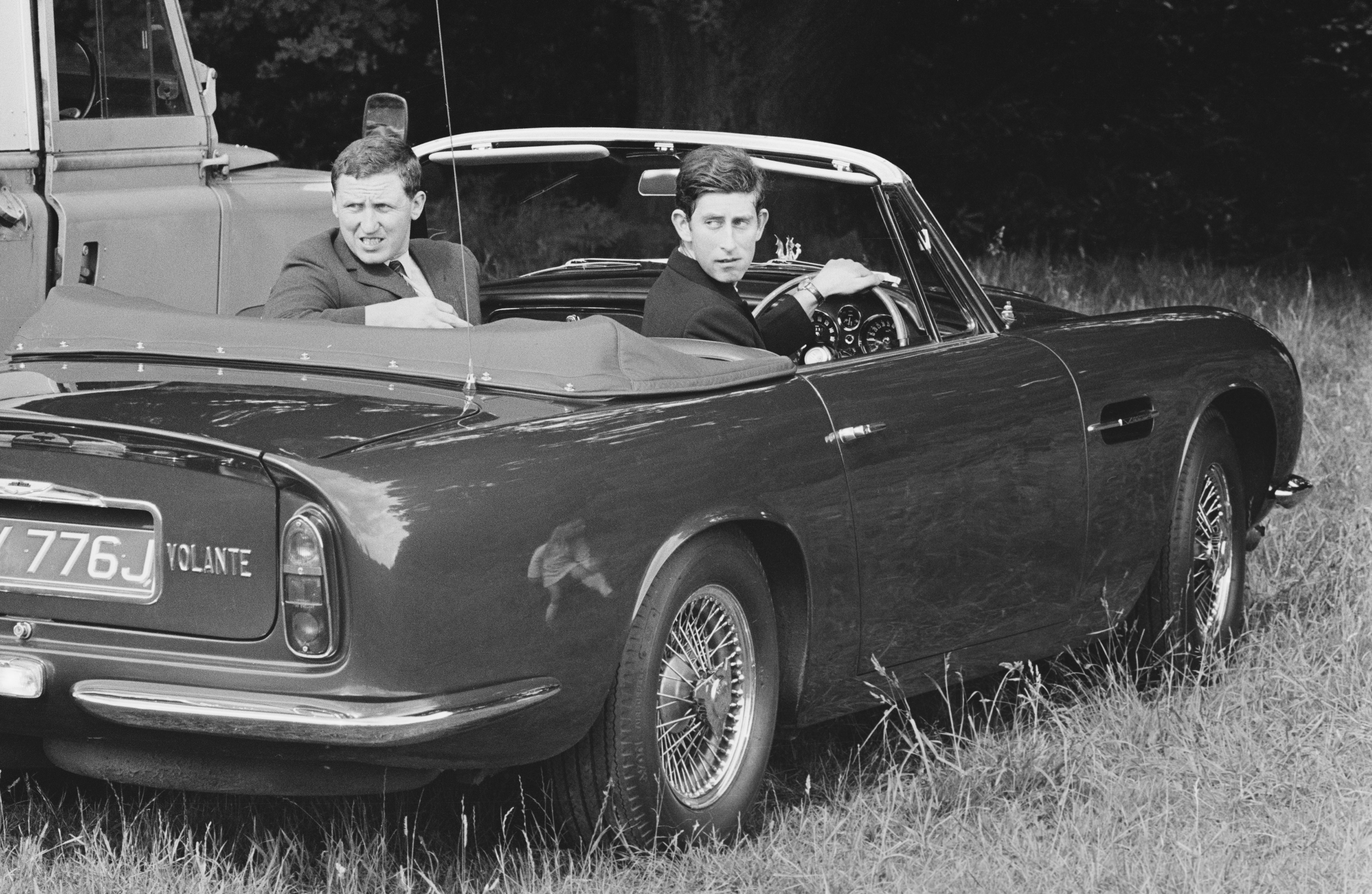 The Prince of Wales and a friend look over their shoulders while reversing out of a parking spot in an Aston Martin Volante