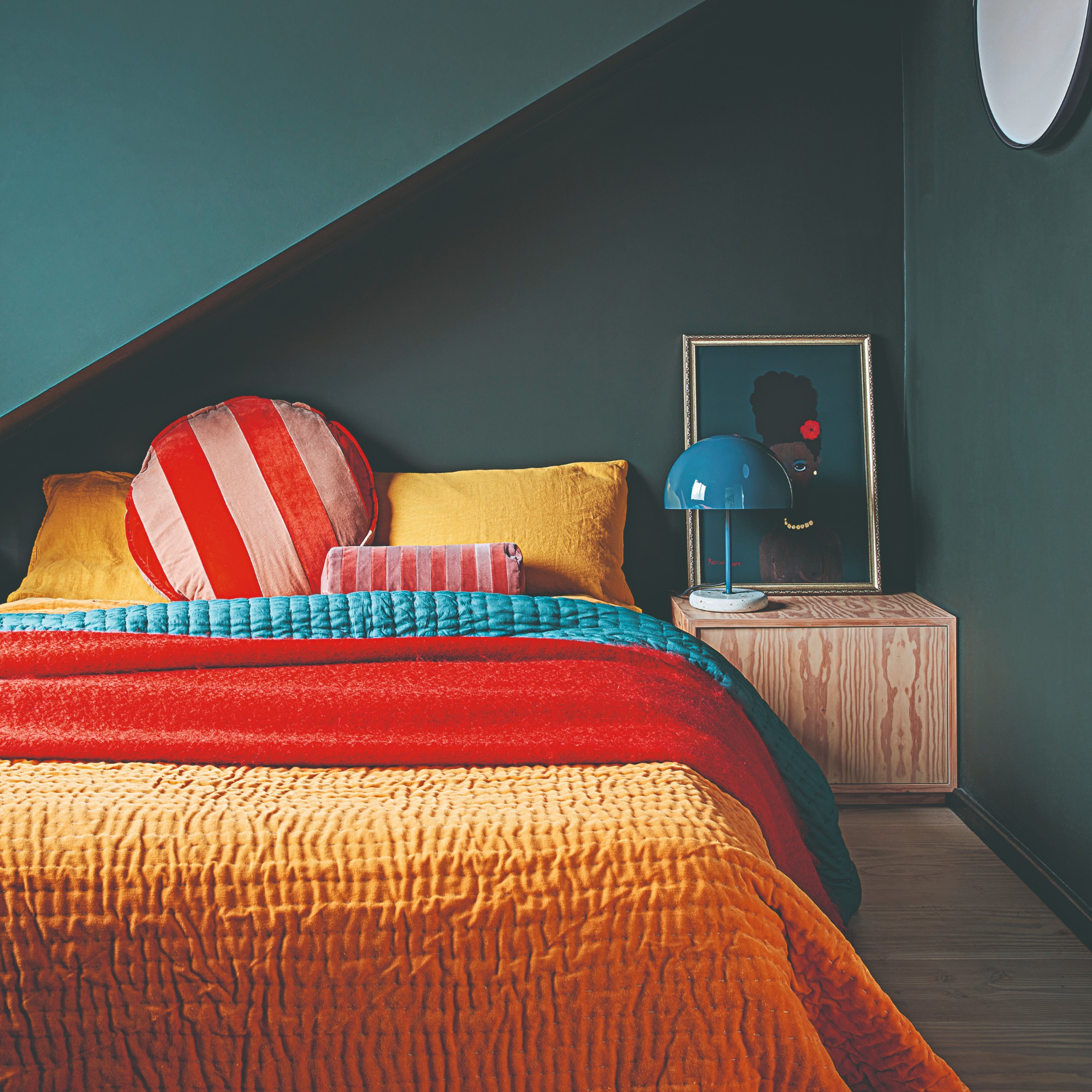 A small bedroom painted in dark teal with a bed dressed in red and yellow bedding