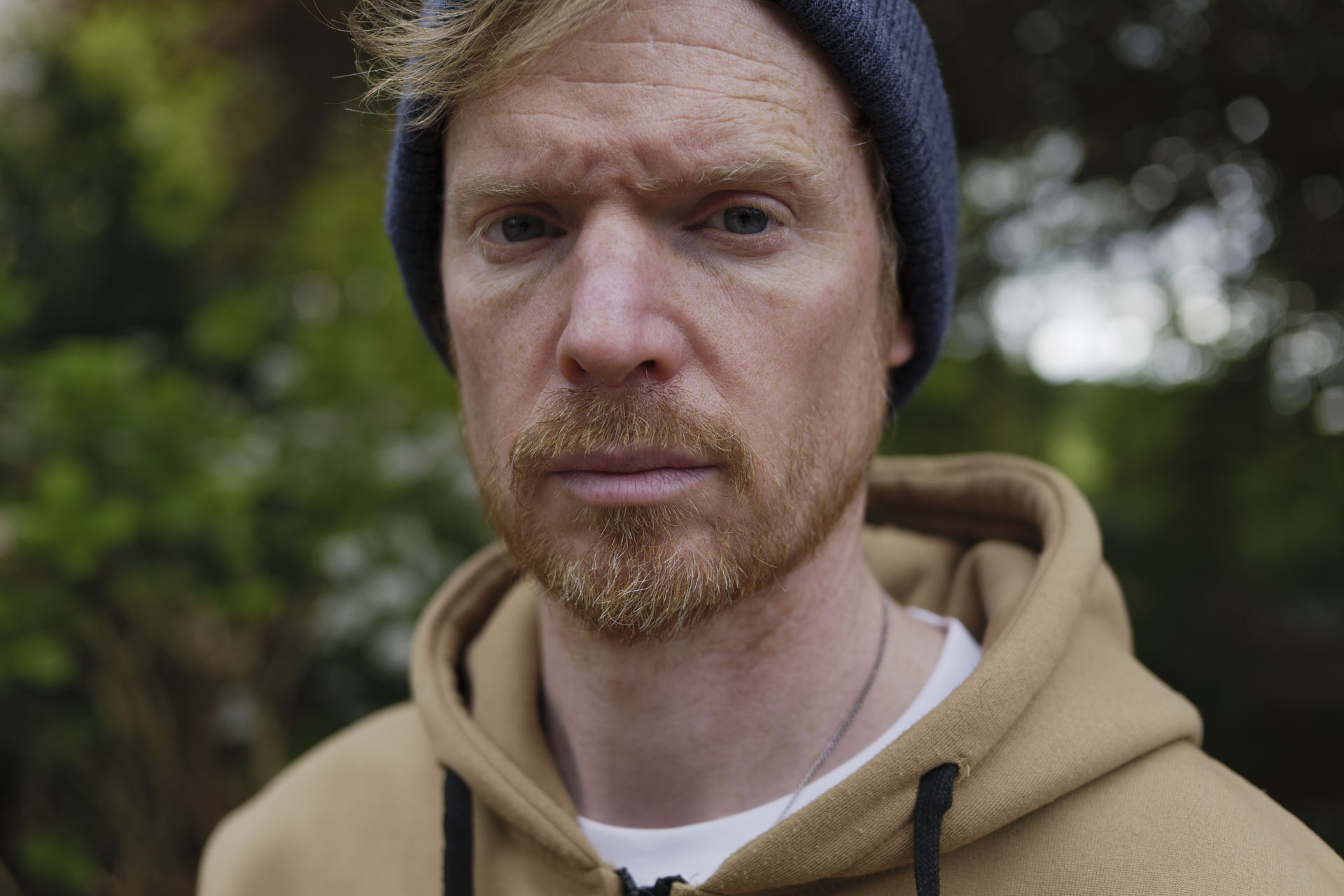 Portrait of a man in a beanie hat and hoodie, the background is lush green bushes and trees with dappled light