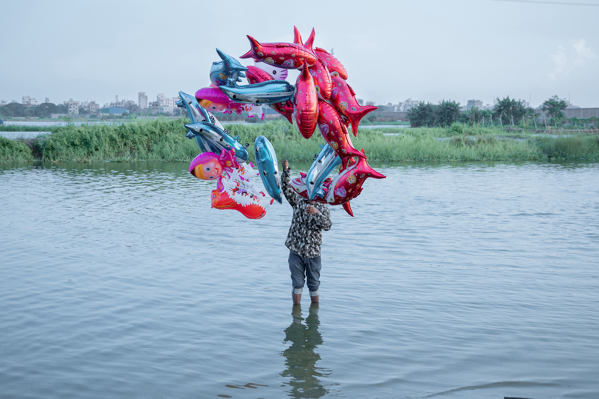 The fish belong in the river, but the river is gone. A man stands in the water, while balloons shaped like dolphins float above, a parody of joy