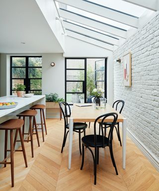 side extension with brick internal wall painted white and white angled glazed roof above, dining table and breakfast bar in shot with wooden floor