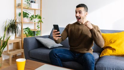 A man celebrates as he looks at his phone while on his sofa.