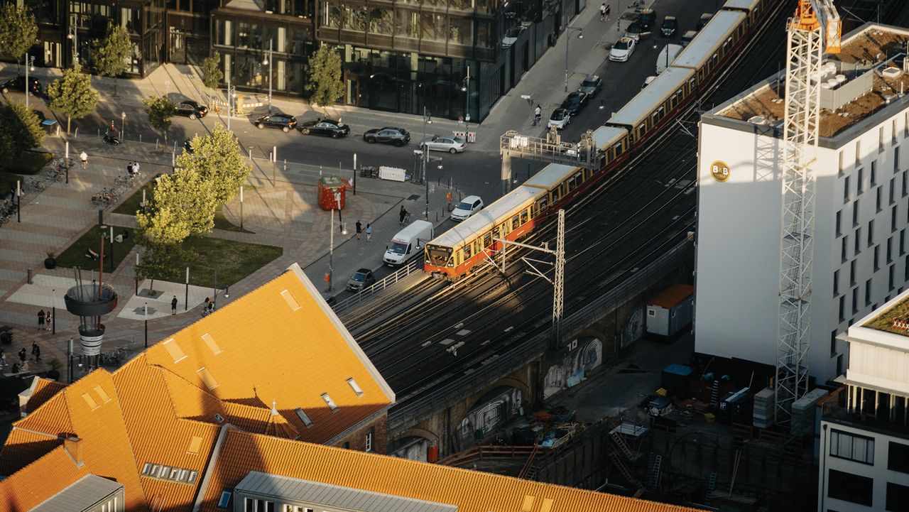 Aerial view of a train on tracks near modern buildings, with cars and pedestrians on a nearby street, showcasing urban life