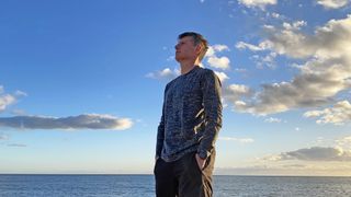 A man stands wearing a Saxx Roast Master Mid-Weight Long Sleeve base layer, the sea and sky behind him