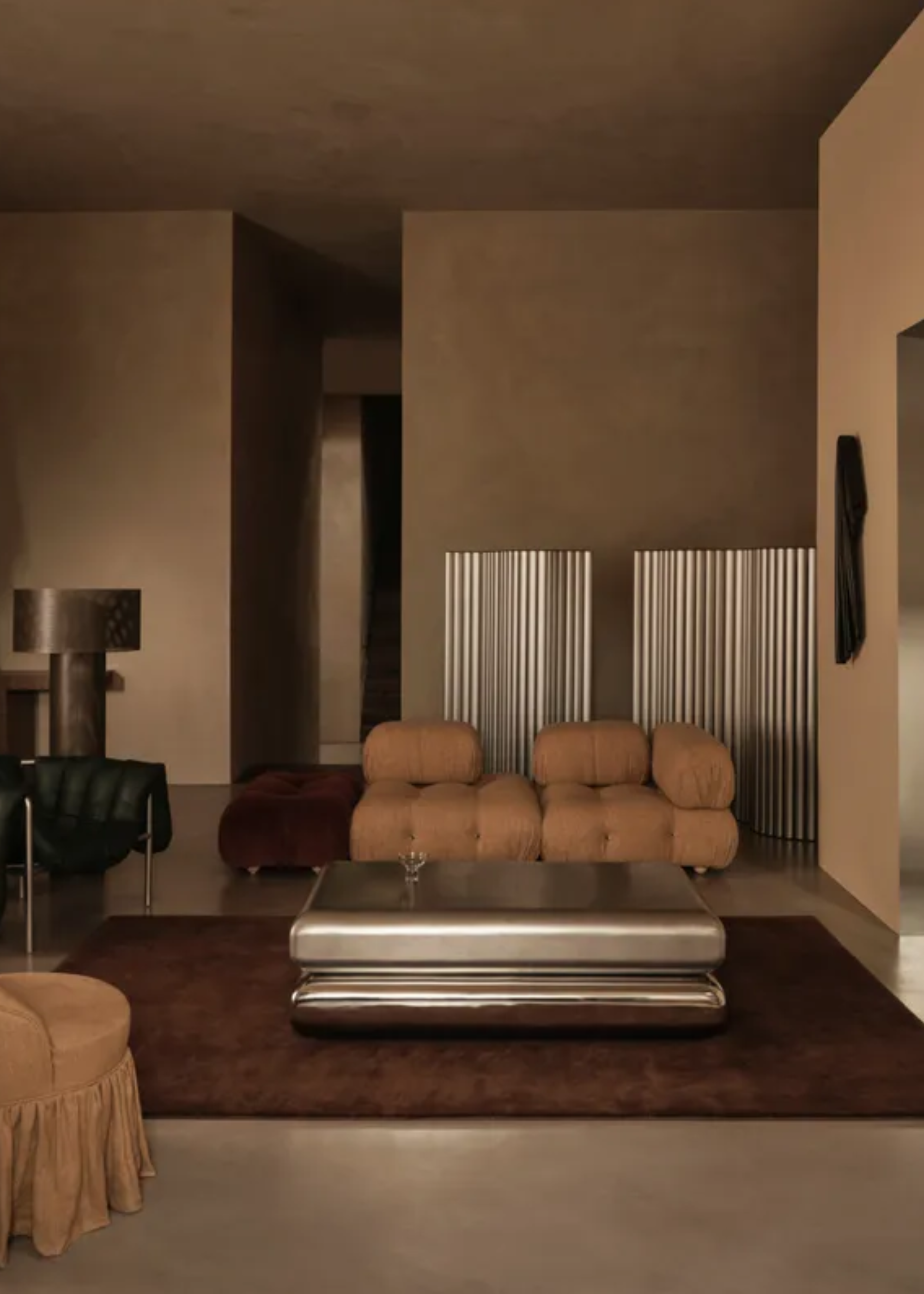 A sandy colored room with chrome room dividers and a coffee table on a brown rug by a beige sofa set with a brown seater and a dark silver lamp