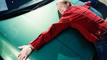 A car-shopping woman hugs the hood of a new car.