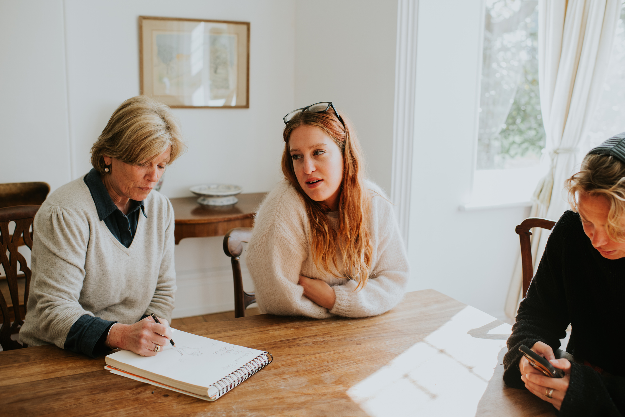 Two younger woman sit around a dining table with an older woman who takes notes on a notepad.