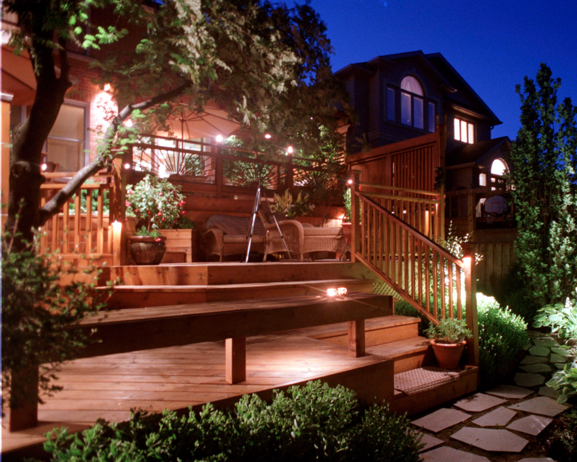 front yard patio area accessed by wooden steps and deck, with lighting, ornamental trees, planting and garden furniture