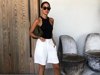 Influencer Tania Sarin poses outside the Proper Hotel in Los Angeles wearing black cat-eye sunglasses, a Bottega Veneta cloud pouch clutch, black racerback tank top, and baggy white linen Bermuda shorts