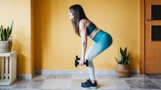 woman in leggings and crop top holding two dumbbells half way through a deadlift. she's sideways to the camera with a yellow wall and a couple of plants behind her 
