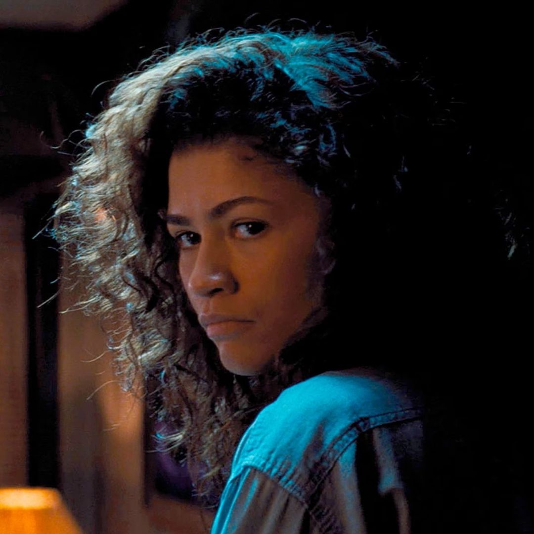 zendaya as rue looking over her shoulder in a dimly lit room in a still from euphoria season 3